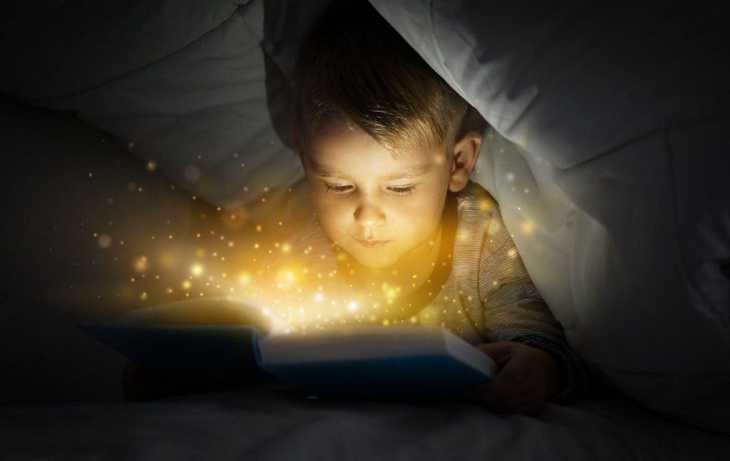 Cute little boy reading magic book in bed under blanket