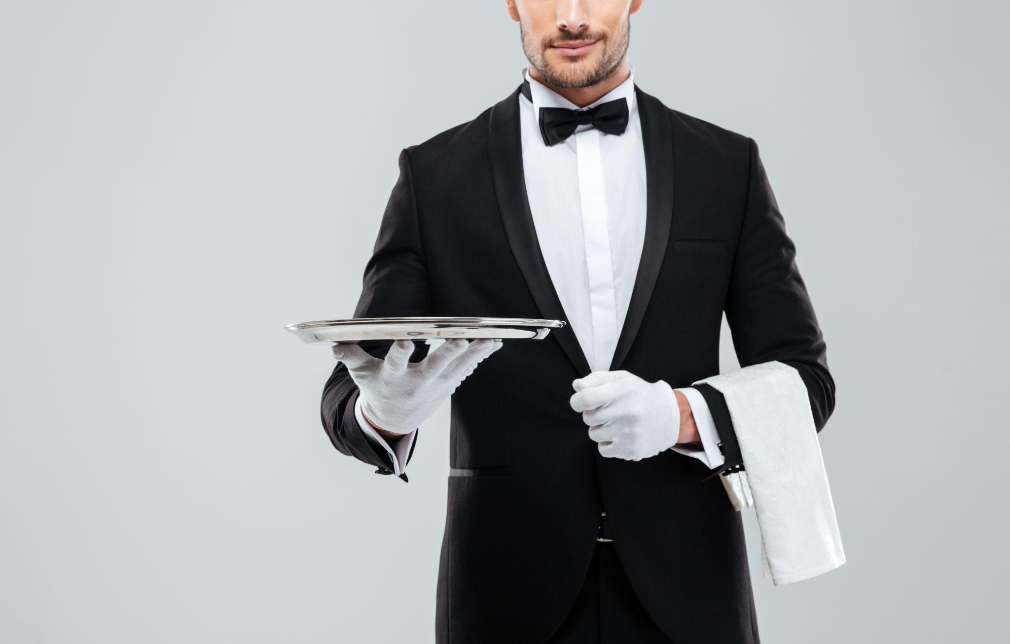 Closeup of young waiter in tuxedo holding metal empty tray and napkin