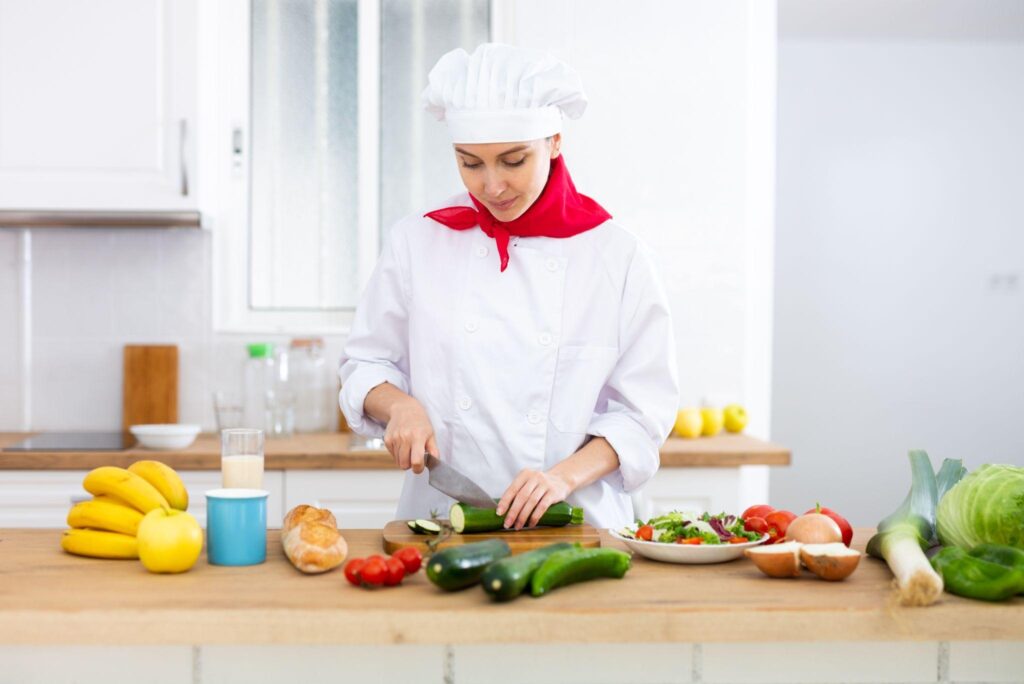 Smiling female chef in white uniform preparing vegetable salad in private kitchen